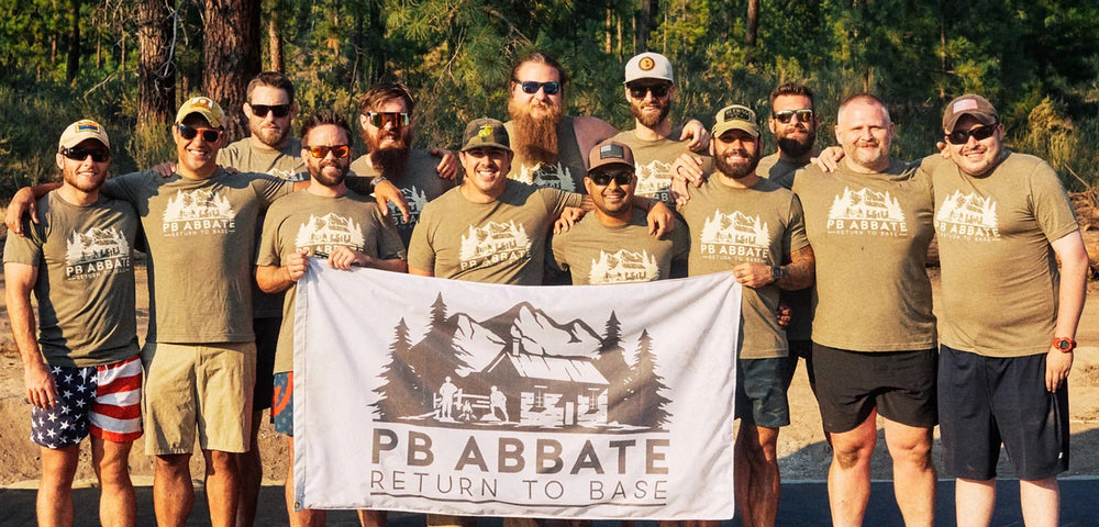 A group of 12 men standing outdoors in a forest clearing, wearing matching olive green t-shirts and holding a white banner that reads 'PB ABBATE RETURN TO BASE' with a cabin graphic.