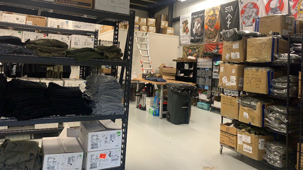 A warehouse storage room with metal shelving units filled with folded clothing, cardboard boxes, and packaged goods. Posters and signs are visible on the wall, and fluorescent lights illuminate the space.